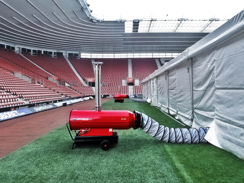Biemmedue Arcotherm EC85 dieselverwarming met schouw en warmeluchtslang naast een tijdelijke tent in een voetbalstadion